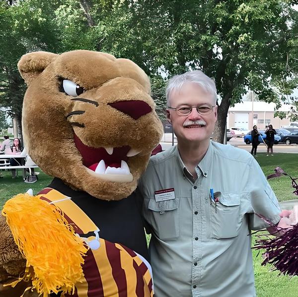 The University of Minnesota Morris Cougar mascot with Steve Burks, a white man with grey hair and mustache. He has glasses and is wearing a green short-sleeved shirt and holding a maroon pompom